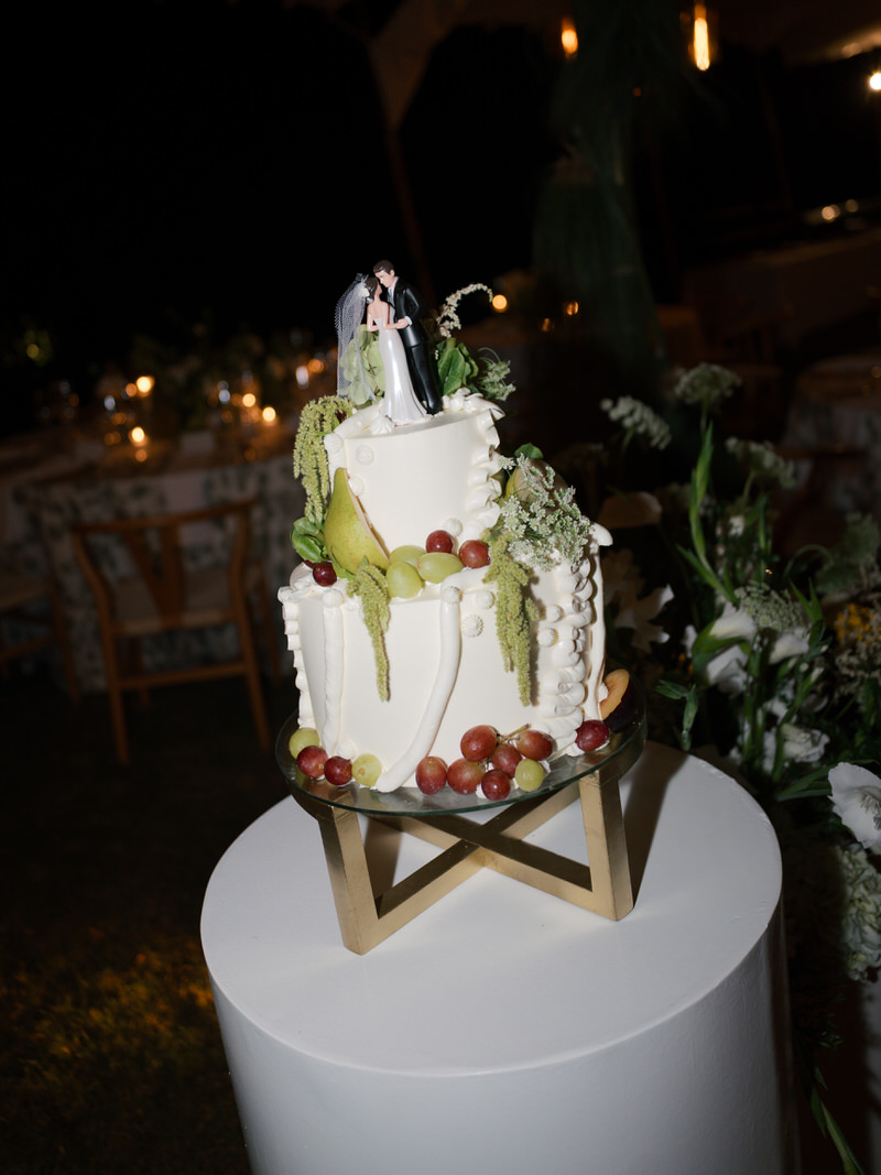 Two-tier white wedding cake decorated with fresh fruit, greenery, and a bride and groom topper at a Costa Rica destination wedding reception